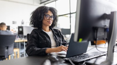 A female employee smiles at her laptop screen. She has curly, brown, shoulder-length hair and wears glasses with black plastic frames.