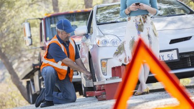 A tow truck driver kneels beside the tire of the disabled vehicle; the driver of the car stands in front of the vehicle, looking at her smartphone.